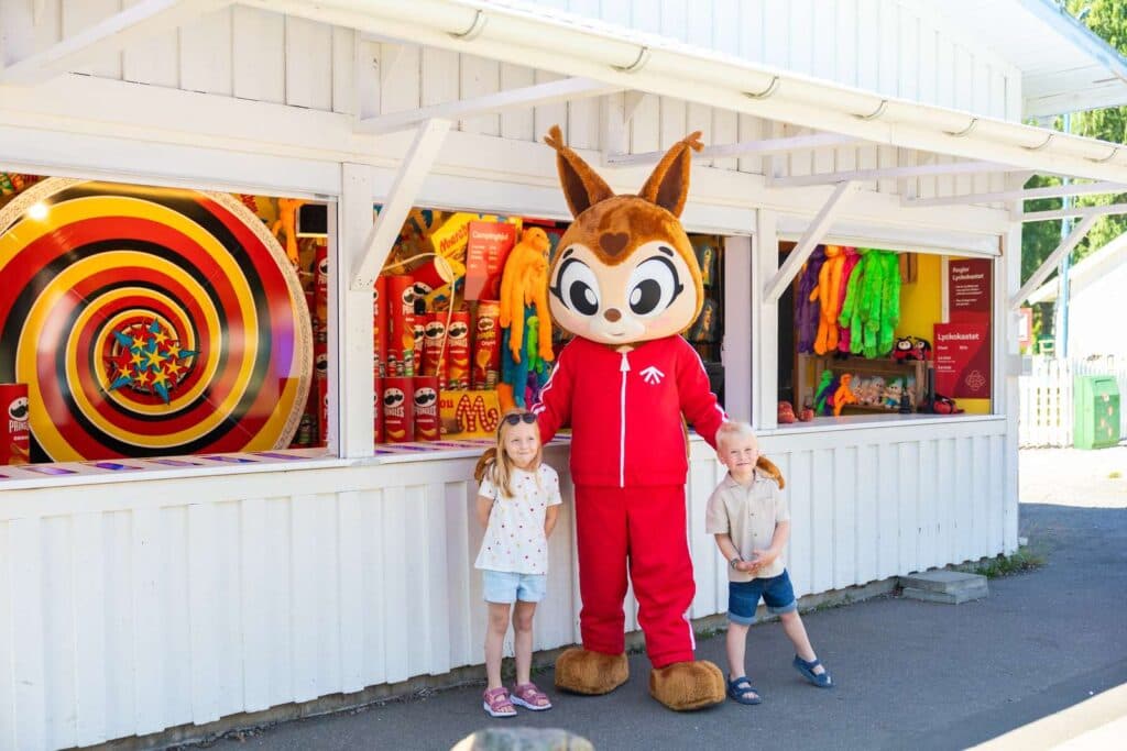 Maskoten Yessi tillsammans med två barn framför lyckohjulet på Skara Sommarland camping